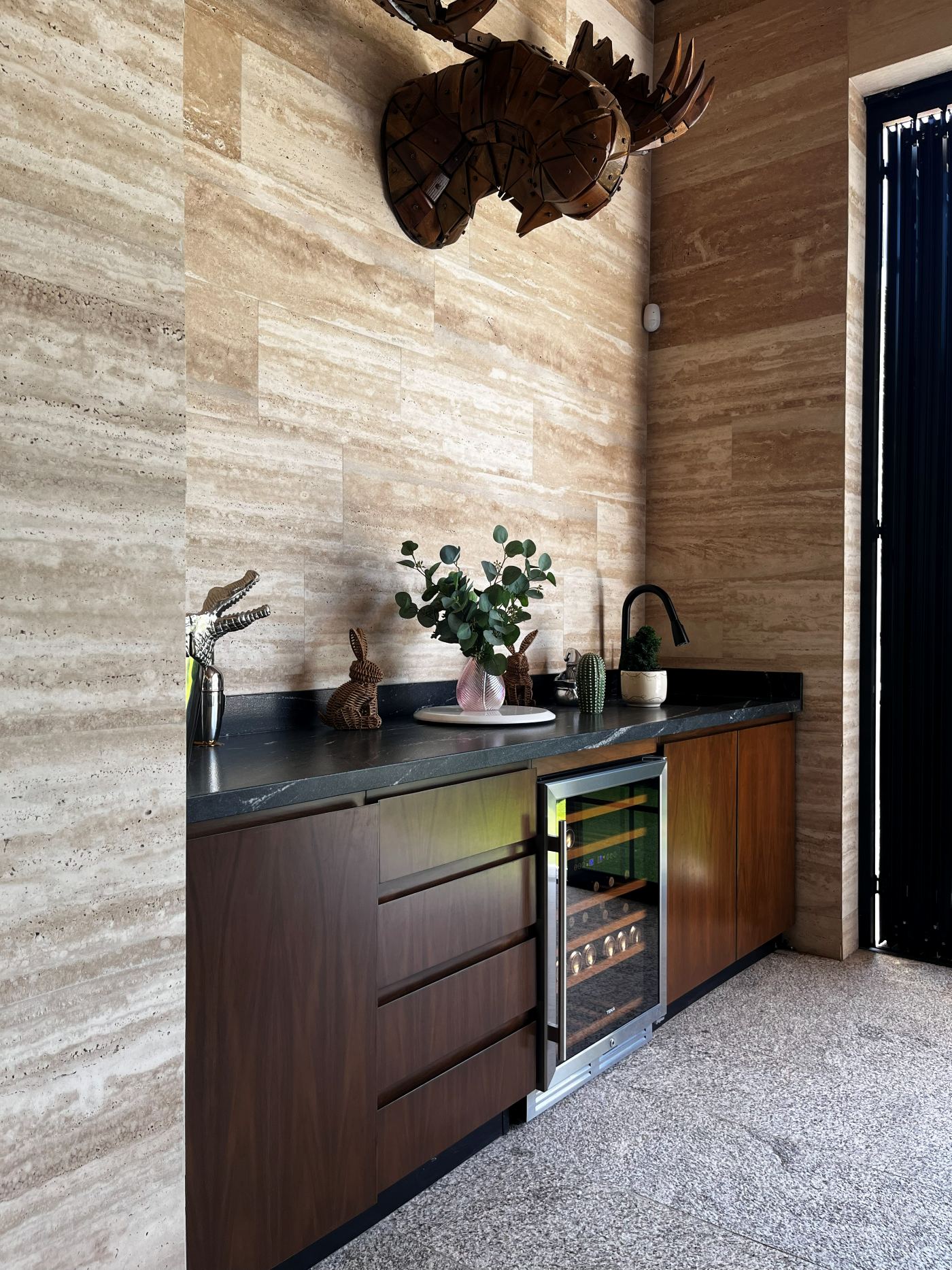 Barra con gabinetes de madera oscura y frigobar bajo un muro de travertino
